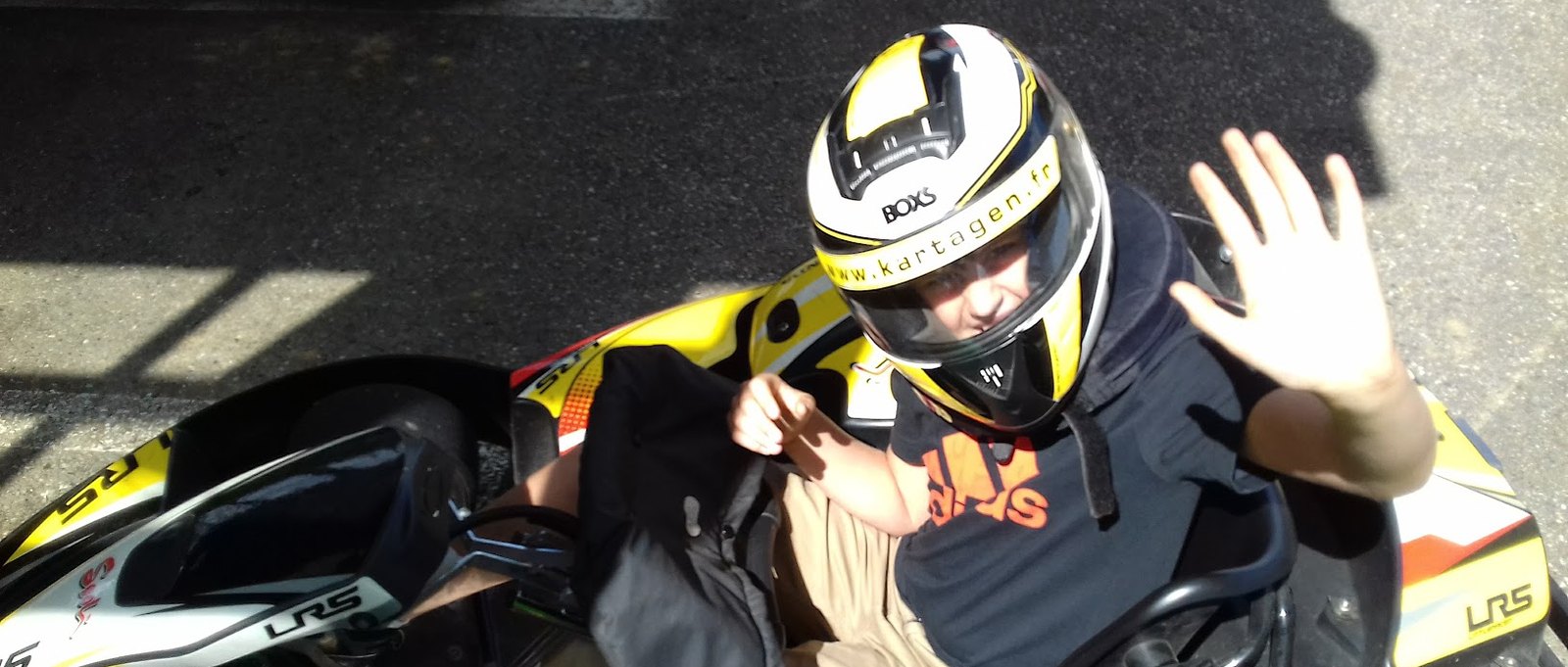A young person in a helmet sitting in a go-kart, waving at the camera